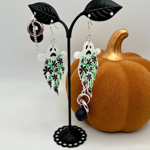 May include: White ghost-shaped earrings with black eyes and mouths, adorned with black and green floral patterns. One earring has a pink ring and black bead attached. The earrings are displayed on a black stand with a pumpkin in the background.