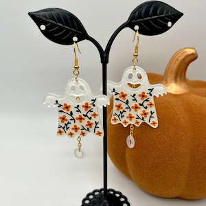 May include: A pair of ghost-shaped earrings with gold-colored hooks. The ghosts are white with orange and black floral patterns. Each earring has a small, clear, oval-shaped charm dangling from the bottom. A pumpkin is in the background.