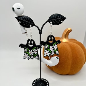May include: Black ghost-shaped earrings with white daisy and green leaf accents. The earrings have a smiling face and are displayed on a black stand with a white background. A decorative orange pumpkin is in the background.