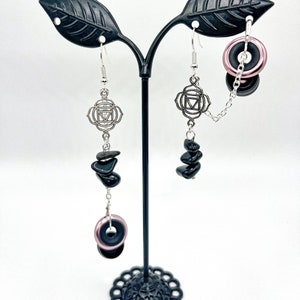 May include: A pair of silver earrings with black stones and a silver charm with a symbol. One earring has a pink and black ring charm.