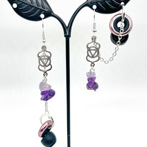 May include: A pair of silver earrings with amethyst stones and a silver charm with a geometric design. The earrings are hanging from a black stand.