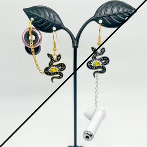 May include: Earrings displayed on a black stand. One earring is a gold hoop with a pink and black plug and a gold chain. The other earring is a black snake design with a yellow sun and a silver chain with a white earbud.