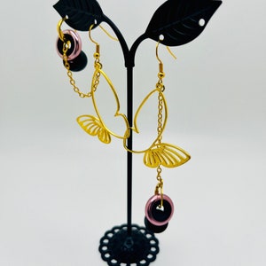 May include: Gold butterfly earrings with dangling pink and black beads. The earrings are on a black stand.