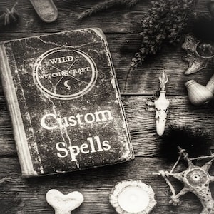 Puede incluir: Un libro negro con el título "WILD WITCH CRAFT" y "Custom Spells" en la portada. El libro está rodeado de varios objetos, incluyendo una pipa, una pluma, una pluma y una estrella de madera.