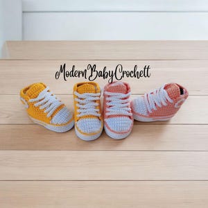 May include: Four handmade crochet baby sneakers arranged in a row. Two shoes are yellow with white laces and white toe caps, and two are peach with white laces and white toe caps. The text Modern Baby Crochett is centered above the shoes.