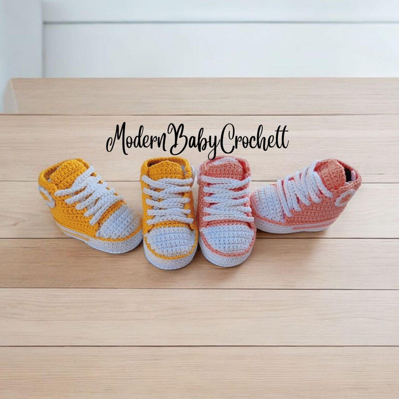 May include: Four handmade crochet baby sneakers arranged in a row. Two shoes are yellow with white laces and white toe caps, and two are peach with white laces and white toe caps. The text Modern Baby Crochett is centered above the shoes.