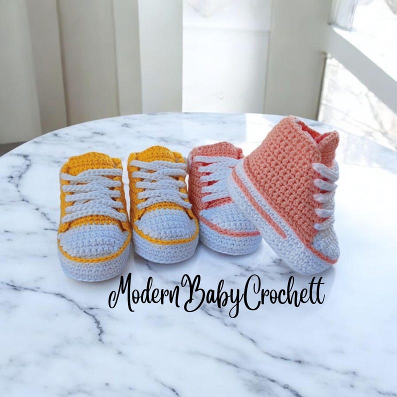May include: Four handmade crochet baby booties designed to resemble sneakers. Two are yellow with white laces and toe caps, while the other two are peach-colored, including one high-top style. The text Modern Baby Crochett is visible.