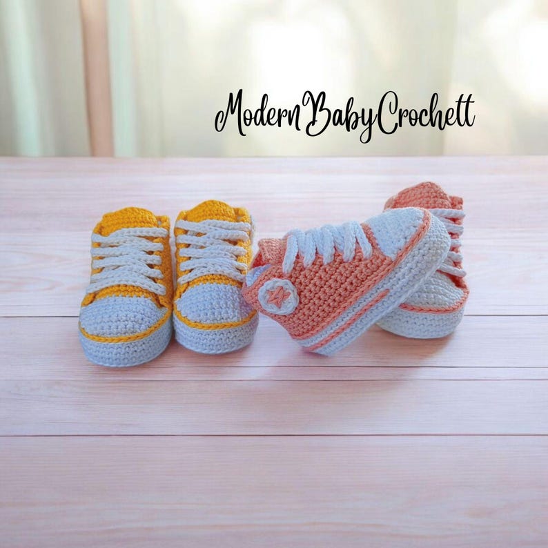 May include: Two pairs of handmade crochet baby sneakers. One pair is bright yellow with white laces and toe caps. The other pair is peach-colored with white laces, white toe caps, and a circular side patch. The text Modern Baby Crochett is visible.
