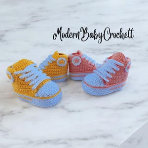 May include: Two pairs of handmade crochet baby sneakers with the text Modern Baby Crochett. One pair is yellow with light blue soles and laces, and the other pair is peach with light blue soles and laces, featuring circular side patches.