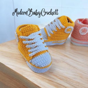 May include: A pair of mustard yellow and light gray crochet baby sneakers with white laces and a circular side patch. The text Modern Baby Crochett is visible at the top. These high-top style shoes feature a textured yarn construction.