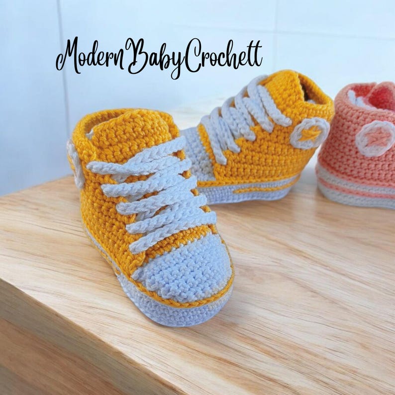 May include: A pair of mustard yellow and light gray crochet baby sneakers with white laces and a circular side patch. The text Modern Baby Crochett is visible at the top. These high-top style shoes feature a textured yarn construction.