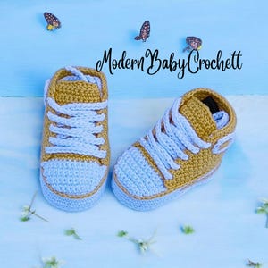 May include: A pair of mustard yellow and white crochet baby sneakers with white laces and a rounded toe cap. The text Modern Baby Crochett is visible above the shoes, accompanied by three small butterfly illustrations on a light blue background.