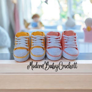 May include: Two pairs of handmade crochet baby sneakers, one pair in mustard yellow and one pair in peach, both featuring white laces and white toe caps. The text Modern Baby Crochett is printed in black on the light wood surface below the shoes.