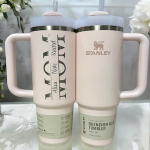 May include: Two pink stainless steel travel mugs with lids and handles. The mug on the left has the text "MOM" and "Alisa Nate Rachel" engraved on it. The mug on the right has the Stanley logo and the text "The Flowstate Quencher H2.0 Tumbler 30 oz / 887 ml All Day Hydration" on it.