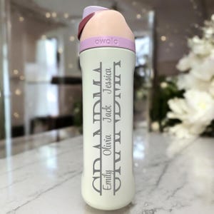 May include: A white insulated water bottle with a light purple lid and a dark red carrying strap. The bottle has the word "owala" printed on the lid and the word "Grandma" and the names "Emily, Olivia, Jack, Jessica" printed vertically on the bottle.