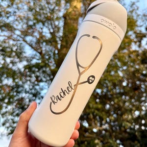 May include: A white Owala water bottle with a grey lid and handle. The bottle has a gold stethoscope design and the name "Rachel" in cursive. The background is blurred with green and brown foliage.