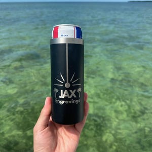 May include: A black stainless steel can cooler with a silver logo that says "JAX Engravings" and a palm tree design. The cooler is holding a silver can with a red, white, and blue label.