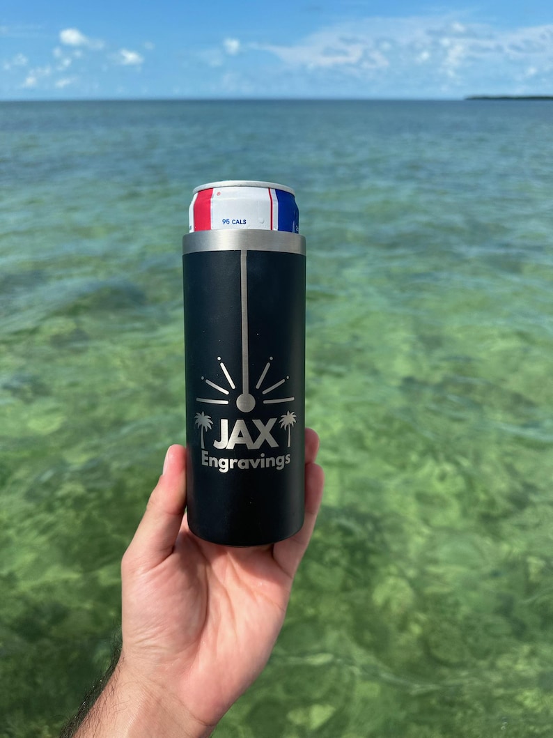 May include: A black stainless steel can cooler with a silver logo that says "JAX Engravings" and a palm tree design. The cooler is holding a silver can with a red, white, and blue label.