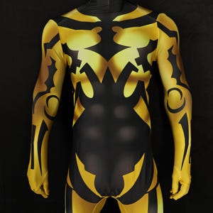 Puede incluir: Un disfraz de cuerpo entero en amarillo y negro, exhibido en un maniquí. El diseño presenta intrincados patrones negros sobre una base amarilla, cubriendo todo el cuerpo, incluidos los brazos y las piernas. El disfraz tiene un cuello alto.