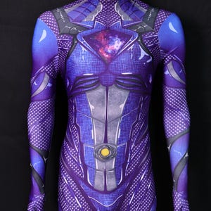 Puede incluir: Mono morado de cuerpo entero con cuello alto, con un diseño detallado con detalles grises y plateados. El área del pecho tiene un diseño en forma de diamante con un patrón de galaxia. Un círculo amarillo está centrado en el abdomen.
