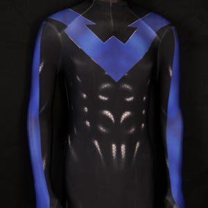 Może przedstawiać: Czarny i niebieski kostium superbohatera. Całkowity strój ciała ma niebieski wzór w szewron na klatce piersiowej i ramionach, z niebieskimi akcentami wzdłuż ramion. Strój ma serię białych, o strukturze plastra miodu detali na tułowiu.