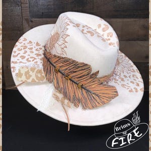 May include: A cream-colored fedora hat with a wide brim, adorned with a large, detailed feather and sunflower and leopard print accents. The hat features the text "Brims of FIRE" in a circular logo.