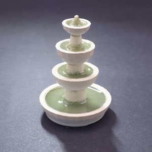 May include: A miniature, tiered fountain with a white exterior and light green water. The fountain has four levels, each with a circular basin. The top level has a small, pointed finial. The fountain is set against a dark gray background.