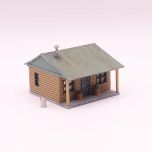 May include: A miniature model of a small cabin with a corrugated metal roof. The cabin has a light brown wooden exterior, a front porch, and a chimney. Two small potted plants sit near the front door. A small metal trash can is next to the cabin.