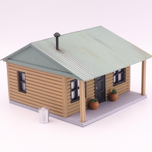 May include: A miniature model of a small house with a light green corrugated metal roof and a porch. The walls are made of horizontal wooden planks. The house has a black door and windows, and two small potted plants on the porch.