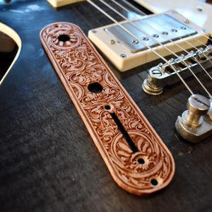 Könnte beinhalten: Eine braune, aufwendig geschnitzte Gitarren-Bedienplatte mit floralen und scrollartigen Designs. Die Platte befindet sich auf einem dunklen Holzgitarrenkorpus, in der Nähe der Saiten und Tonabnehmer. Die Platte hat drei Löcher und einen langen Schlitz.