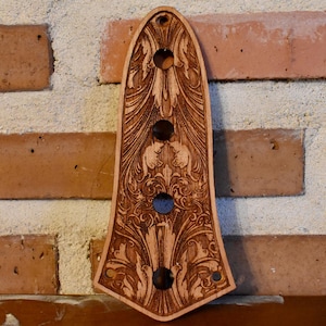 Könnte beinhalten: Eine hellbraune Gitarrenkopfplatte aus Holz mit aufwendigen floralen und scrollartigen Schnitzereien. Sie hat drei dunkelbraune Knöpfe und drei runde Löcher für Stimmwirbel. Die Platte ist an einer Backsteinmauer befestigt.