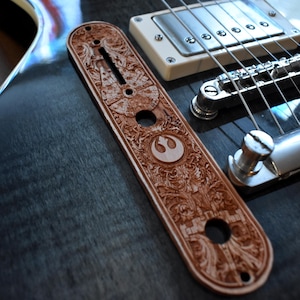 Puede incluir: Una placa de mástil de guitarra marrón, intrincadamente tallada, con un símbolo de la Alianza Rebelde de Star Wars. La placa es de madera y está colocada en una guitarra eléctrica azul oscuro, mostrando patrones florales y geométricos detallados.