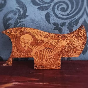Può includere: Un battipenna per chitarra in legno marrone con un intricato design inciso al laser raffigurante un teschio umano e una cassa toracica su uno sfondo di dettagliati motivi floreali. L'oggetto ha una forma curva e asimmetrica con fori di fissaggio.