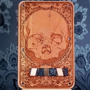 Puede incluir: Panel decorativo de madera con un detallado grabado de calavera dentro de un marco ornamental de inspiración floral. El panel tiene forma rectangular con un tono de madera más claro y una base decorativa. El diseño está intrincadamente tallado, mostrando un tema gótico o macabro.