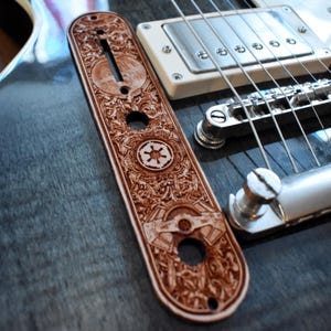 Könnte beinhalten: Eine braune, lasergravierte Gitarren-Bedienplatte mit aufwendigen floralen und geometrischen Mustern, einschließlich eines kreisförmigen Emblems. Die Platte liegt neben einer blau-grauen E-Gitarre mit silbernen Saiten und Hardware.