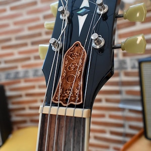 Puede incluir: Primer plano de un clavijero de guitarra negro con cuerdas y clavijas. Una pieza decorativa de madera con símbolos tallados y la palabra "Zeppelin" está unida al clavijero. El fondo es una pared de ladrillos borrosa.