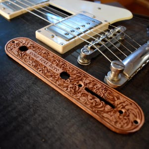 Könnte beinhalten: Eine braune, gravierte Gitarren-Bedienplatte mit dem Text "your text here" wird neben einer Gitarre angezeigt. Die Platte hat ein verziertes Blumendesign und ist aus Holz gefertigt. Die Gitarre hat einen cremefarbenen Korpus und silberne Hardware.