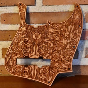 May include: A light brown wooden bass guitar pickguard, intricately carved with a floral pattern. The design features detailed leaves and swirling patterns. The pickguard has pre-drilled holes for mounting, and is made of wood.