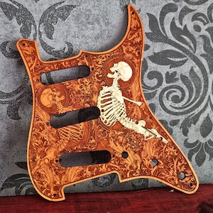 Könnte beinhalten: Ein hölzernes Gitarren-Schlagbrett mit einem detaillierten, lasergravierten floralen Rankenmuster und einer Skelettfigur. Das Design umfasst drei rechteckige Tonabnehmer-Aussparungen, mehrere kreisförmige Schraublöcher und eine hellbraune Oberfläche.