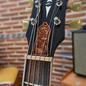 Puede incluir: Primer plano del clavijero de una guitarra eléctrica negra con una placa decorativa de color cobre con un diseño floral ornamentado. El clavijero tiene clavijas de afinación color crema y una cejuela blanca. El fondo es una pared de ladrillos borrosa.