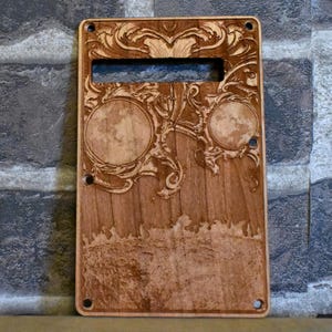 Può includere: Un pickguard per chitarra in legno con un intricato design tagliato a laser che presenta due lune e motivi a spirale. Il pickguard ha una finitura in legno marrone e una forma rettangolare.