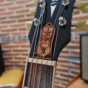 May include: Close-up of a black guitar headstock with tuning pegs and strings. A decorative wooden inlay features a detailed design with a bat-like creature. The guitar's fretboard and headstock are visible against a blurred brick background.