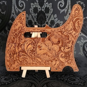 Può includere: Un battipenna per chitarra in legno marrone chiaro con intricati motivi floreali e a volute incisi al laser. Il centro presenta un'illustrazione dettagliata di due scheletri con aureole, circondati da decorazioni e fori di montaggio.
