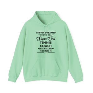 May include: A light green hoodie with a front pocket and a drawstring hood. The front of the hoodie features the text "I NEVER DREAMED I WOULD BE A Super Cool TENNIS COACH BUT HERE I AM KILLING IT!" in black.