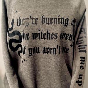 Puede incluir: Sudadera gris con texto gótico negro: "they're burning all the witches even if you aren't me." Un gráfico de serpiente negra está en la parte delantera. La manga derecha tiene el texto "light me up".