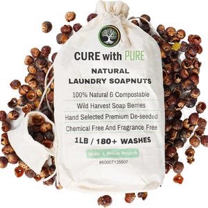 May include: A white drawstring bag labeled "CURE with PURE NATURAL LAUNDRY SOAPNUTS" with a tree logo. The bag is surrounded by brown soap nuts. Text on the bag includes "100% Natural & Compostable" and "1LB / 180+ WASHES."