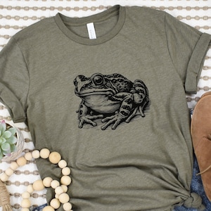 May include: Olive green t-shirt featuring a detailed black and white illustration of a frog. The frog is depicted in a realistic style, with intricate details. The shirt is styled with rolled-up sleeves and a knot at the bottom.