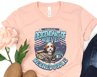 May include: A light pink t-shirt featuring a graphic of a Bernedoodle dog wearing a sweater, with the text "Descendant of Wolves." The design includes a dog bed and food bowl. The shirt is paired with blue jeans and brown boots.