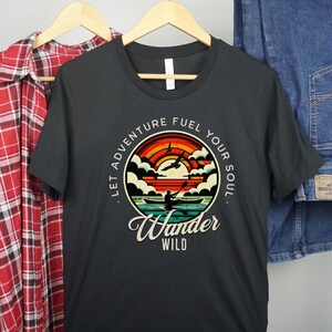 May include: Black t-shirt with a vintage-style graphic of a sunset, clouds, and a person kayaking. The text reads "Let Adventure Fuel Your Soul. Wander Wild."