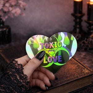 May include: A heart-shaped holographic sticker with the words "TOXIC LOVE" in a colorful, stylized font. The sticker has a biohazard symbol design with green and purple accents. The sticker is held over a dark, textured surface.